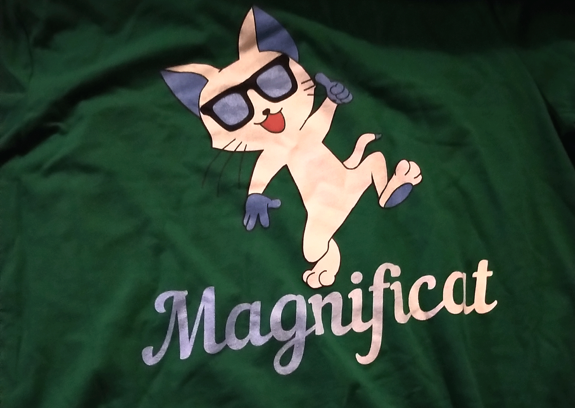 "Green t-shirt with a magnificent looking cat, wearing sunglasses and dancing, and the word Magnificat underneath"